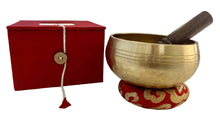 Load image into Gallery viewer, Brass singing bowl with wooden striker on a red cushion, next to a red box with a button, on a white surface.
