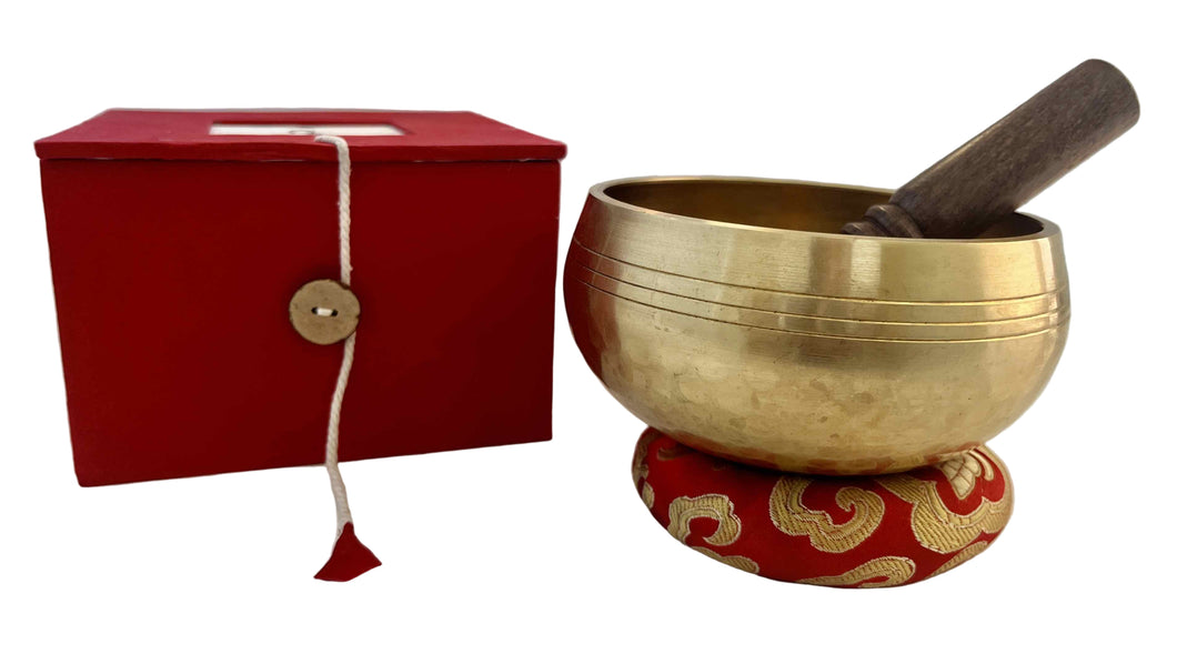 Brass singing bowl with wooden striker on a red cushion, next to a red box with a button, on a white surface.